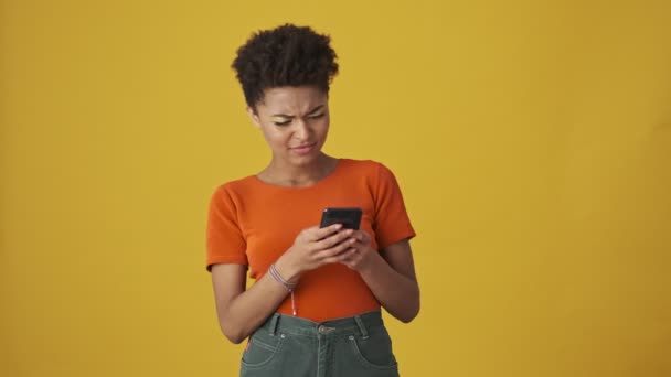 スマートフォンにメッセージを書き 黄色の背景に不満を持つ魅力的なアフリカの女性を喜ばせてください — ストック動画