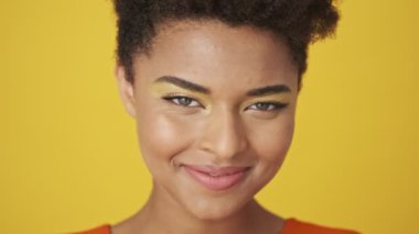 Gülümseyen, neşeli, çekici Afrikalı kadının flört edişini ve sarı arka planda kameraya bakışını yakından izleyin.