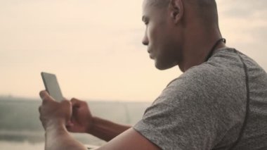 Ciddi bir Afrikalı genç sporcunun yan görüntüsü sabah köprünün dışında akıllı telefonunu kullanıyor.