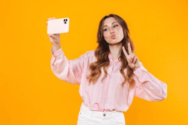 Cep telefonundan selfie çeken ve sarı arka planda izole edilmiş barış işareti yapan hoş bir genç kadın.