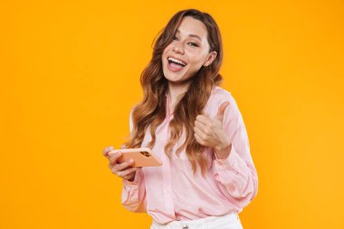 Başparmağını gösteren ve sarı arka planda izole edilmiş cep telefonu kullanan sevimli, neşeli bir kadın görüntüsü