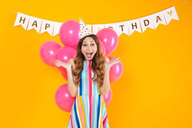 Parti konisi içinde, sarı arka planda izole edilmiş pembe balonlarla poz veren heyecanlı tatlı bir kadın resmi.