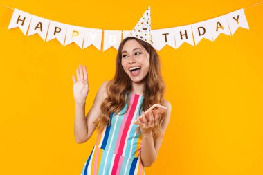 Parti konisinde cep telefonu kullanan ve sarı arka planda el sallayan neşeli sevimli bir kadın görüntüsü