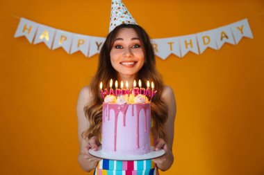 Parti konisinde, sarı arka planda izole edilmiş mumlarla doğum günü turtası gösteren mutlu genç bir kadın resmi.