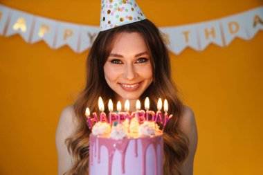 Parti konisinde, sarı arka planda izole edilmiş mumlarla doğum günü turtası gösteren mutlu genç bir kadın resmi.