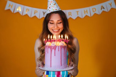 Parti konisinde, sarı arka planda izole edilmiş mumlarla doğum günü turtası gösteren mutlu genç bir kadın resmi.