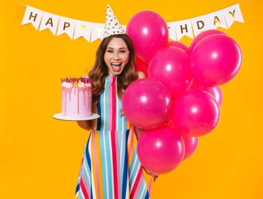 Parti konisi içinde pembe balonlar ve sarı arka planda izole edilmiş doğum günü turtasıyla poz veren heyecanlı tatlı bir kadın resmi.