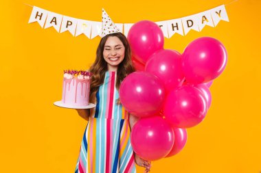 Parti konisi içinde pembe balonlar ve sarı arka planda izole edilmiş doğum günü turtasıyla poz veren heyecanlı tatlı bir kadın resmi.