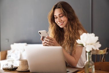 Mutfak masasında oturan, cep telefonu kullanan güzel, genç bir kız.