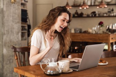 Sevimli neşeli genç kız mutfak masasında oturuyor, video görüşmesi yapıyor, el sallıyor.