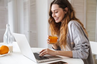 Mutfak masasında oturmuş laptopla çalışıp meyve suyu içerken oldukça neşeli genç bir kadın kahvaltı yapıyor.