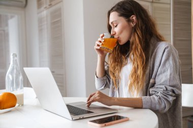 Mutfak masasında oturmuş laptopla çalışıp meyve suyu içerken oldukça neşeli genç bir kadın kahvaltı yapıyor.