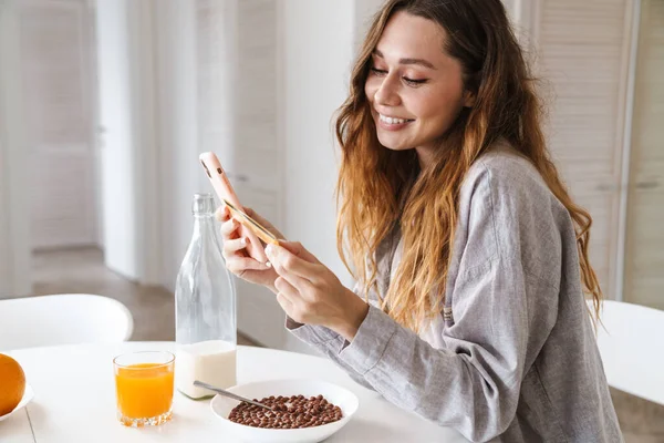 Довольно Веселая Молодая Женщина Завтракает Сидя Кухонным Столом Используя Мобильный — стоковое фото