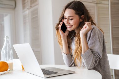 Parlak mutfakta dizüstü bilgisayarla çalışırken gülümseyen ve telefonla konuşan kızıl saçlı kadının portresi