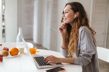 Parlak mutfakta dizüstü bilgisayarla çalışırken gülümseyen ve kulaklık kullanan kızıl saçlı kadının portresi.