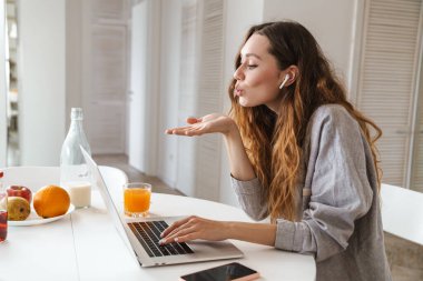 Kulaklıklı mutlu bir kadının portresi. Parlak mutfakta dizüstü bilgisayarla öpüşürken hava öpücüğü veriyor.