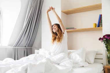 Güzel, mutlu, genç bir kadın sabahları yatakta oturuyor ve ellerini uzatıyor.