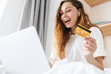 Portrait of cheerful woman with earphone laughing and holding credit card while using laptop in bed clipart