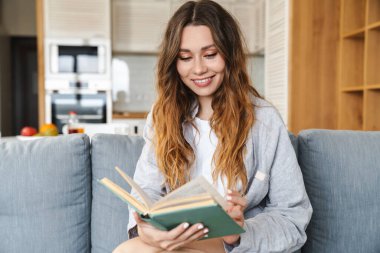 Evdeki kanepede otururken gülümseyen ve kitap okuyan neşeli genç bir kadının fotoğrafı.