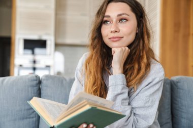 Evdeki kanepede oturmuş kitap okuyan mutlu genç bir kadının fotoğrafı.