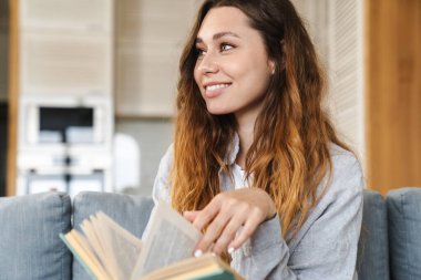 Evdeki kanepede otururken gülümseyen ve kitap okuyan neşeli genç bir kadının fotoğrafı.