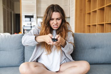 Kızgın genç bir kadın evdeki kanepede dinleniyor, elinde uzaktan kumandayla.