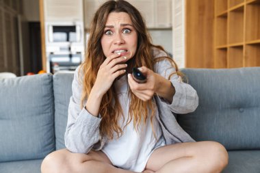 Şok olmuş genç bir kadın evdeki kanepede dinleniyor, elinde uzaktan kumandayla.