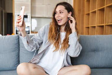 Neşeli genç bir kadın evdeki koltukta dinleniyor, cep telefonu ve kulaklıkla müzik dinliyor.