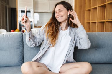 Neşeli genç bir kadın evdeki koltukta dinleniyor, cep telefonu ve kulaklıkla müzik dinliyor.