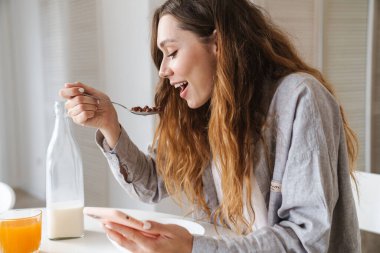 Beyaz mutfakta kahvaltı yaparken gülümseyen ve akıllı telefon kullanan güzel bir kadının fotoğrafı.