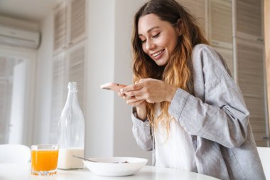Beyaz mutfakta kahvaltı ederken akıllı telefondan fotoğraf çeken neşeli güzel bir kadın resmi.
