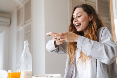 Beyaz mutfakta kahvaltı ederken akıllı telefondan fotoğraf çeken neşeli güzel bir kadın resmi.