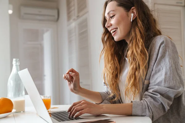 Parlak mutfakta dizüstü bilgisayarla çalışırken gülümseyen ve kulaklık kullanan kızıl saçlı kadının portresi.