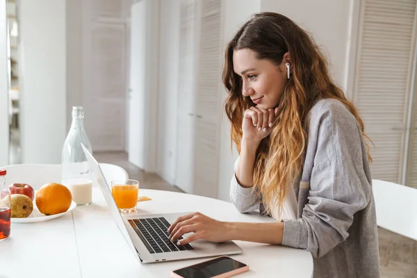 Parlak mutfakta dizüstü bilgisayarla çalışırken gülümseyen ve kulaklık kullanan kızıl saçlı kadının portresi.