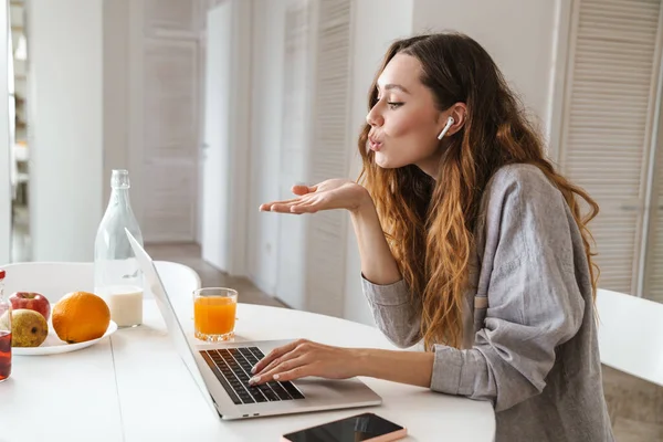 Kulaklıklı mutlu bir kadının portresi. Parlak mutfakta dizüstü bilgisayarla öpüşürken hava öpücüğü veriyor.