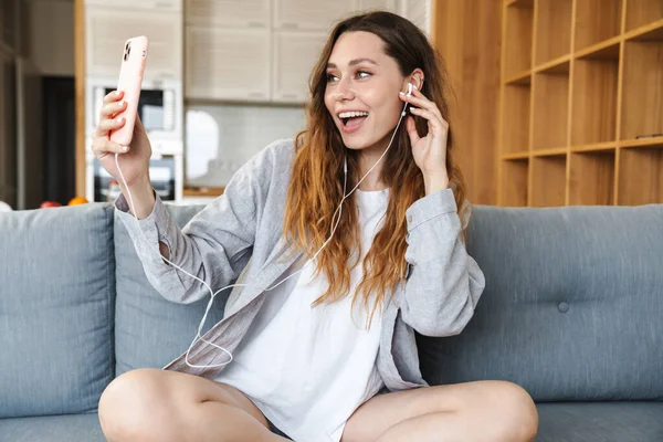 Neşeli genç bir kadın evdeki koltukta dinleniyor, cep telefonu ve kulaklıkla müzik dinliyor.