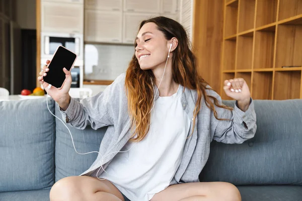快乐的年轻女人在家里的沙发上休息 用手机和耳机听音乐 — 图库照片