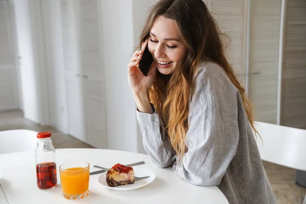 白いキッチンで朝食を食べながら携帯電話で笑ったり話したりする面白い女性の写真 — ストック写真