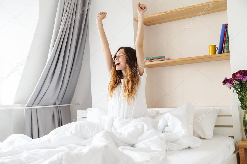 Lovely happy young woman sitting in bed in the morning, stretching hands