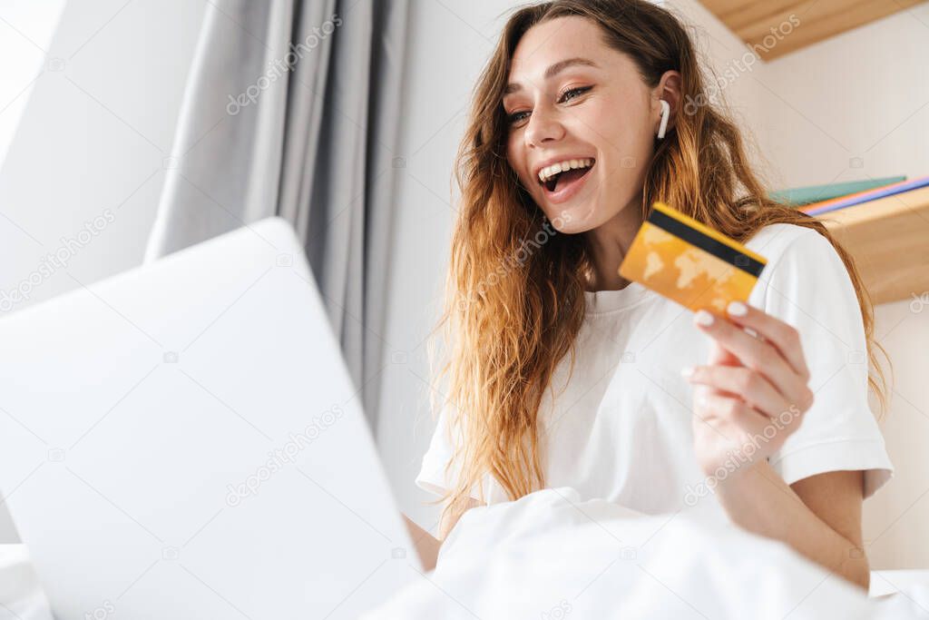 Portrait of cheerful woman with earphone laughing and holding credit card while using laptop in bed