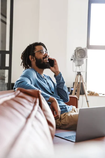 Handsome smiling young african man sitting on a leather couch at home, talking on mobile phone, using laptop computer