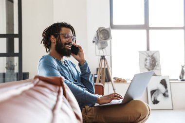 Yakışıklı, gülümseyen Afrikalı bir adam evdeki deri koltukta oturuyor, cep telefonuyla konuşuyor, dizüstü bilgisayar kullanıyor.