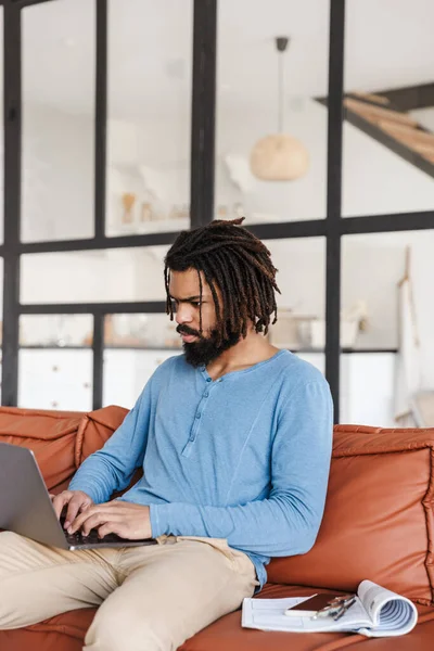 Handsome young african man sitting on a leather couch at home, using laptop computer