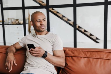 Afro-Amerikan bir adamın oturma odasındaki koltukta otururken cep telefonu kullanması.
