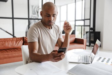 Ciddi bir Afro-Amerikalı adamın oturma odasındaki masada dizüstü bilgisayarla çalışırken cep telefonu kullanmasının fotoğrafı.