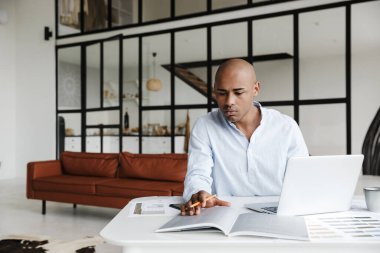 Ciddi bir Afrikalı Amerikalının oturma odasında otururken dizüstü bilgisayarla çalışmasının fotoğrafı.