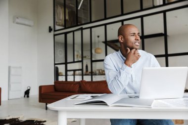 Oturma odasında otururken bilgisayarla çalışan gülümseyen Afrikalı Amerikalı adamın fotoğrafı.