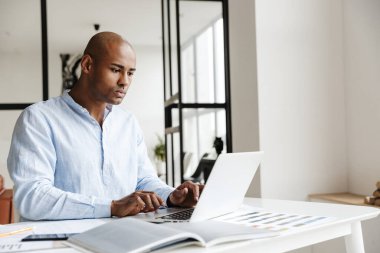 Konsantre olmuş Afro-Amerikan adamın oturma odasında otururken dizüstü bilgisayarla çalıştığı fotoğraf.