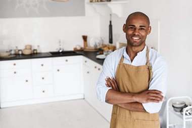 Önlüklü neşeli Afro-Amerikan adamın fotoğrafı. Mutfakta kollarını kavuşturmuş poz verirken gülümsüyor.