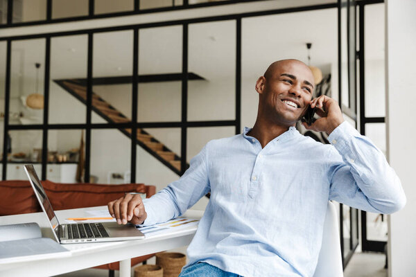 Фото радостного африканского американца, разговаривающего по мобильному телефону во время работы с ноутбуком в гостиной